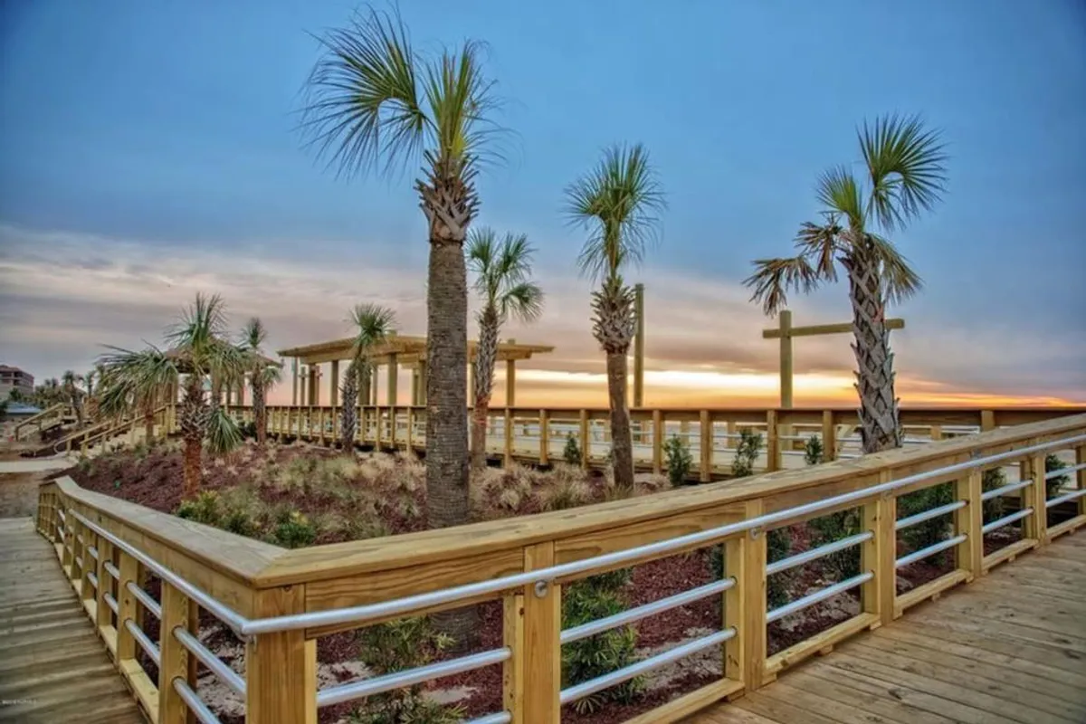 A serene shot of a wooden walkway flanked by palm trees and leading toward the horizon, with a stunning sunset sky. The tranquil atmosphere invites visitors to connect with nature and unwind while taking in the coastal landscape.