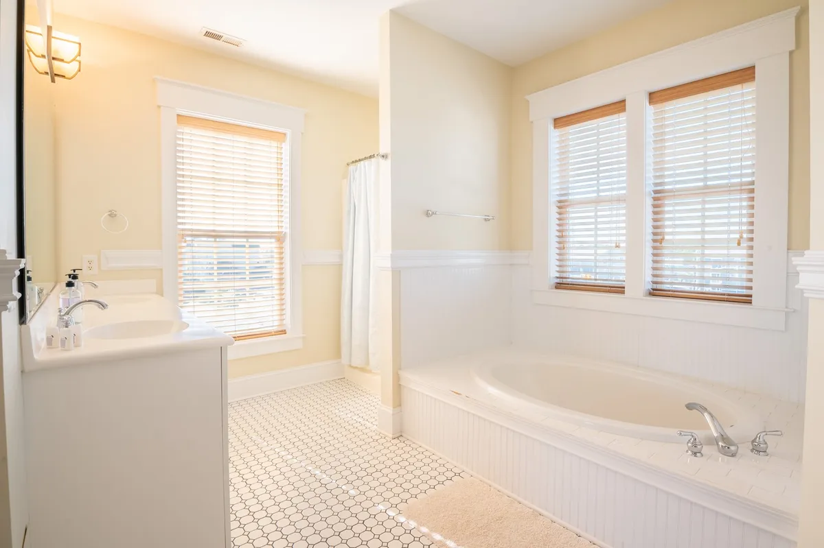 A bright, spa-like ensuite bathroom with a large soaking tub, double windows covered by blinds, and a vanity with a sink. The atmosphere is calm and inviting, ideal for self-care routines and relaxation after a day of adventure.