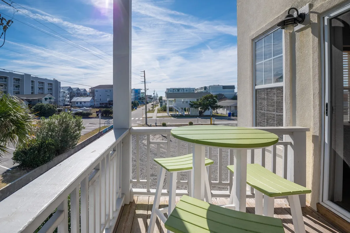 A cozy green table and chairs rest on a balcony or porch with a view of the street and palm trees, creating a welcoming outdoor space for morning coffee or relaxing with a pet.
