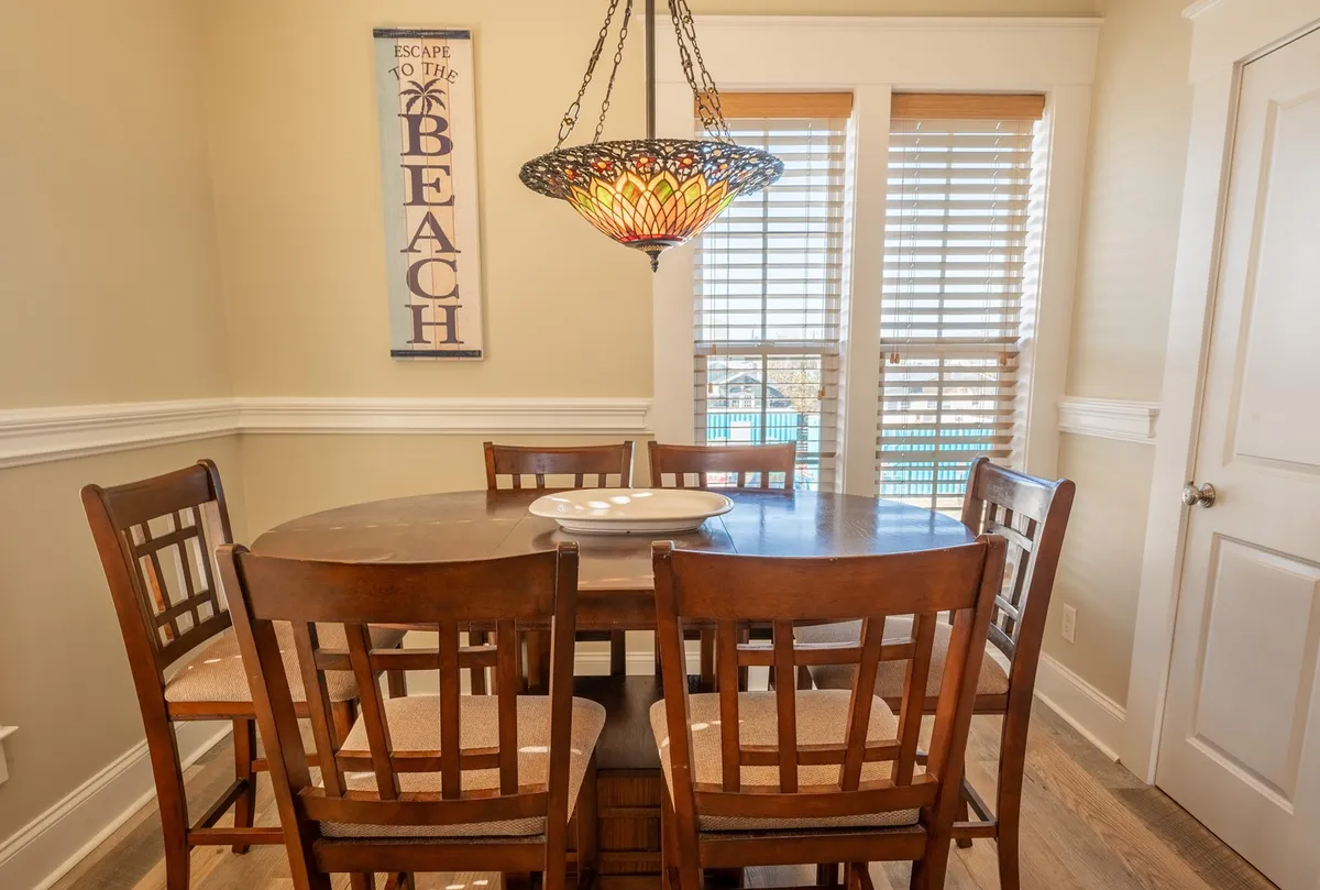 A cheerful and airy dining space with a large table, a colorful stained-glass chandelier, and a sign welcoming guests to escape to the beach. The windows allow in natural light, and the room is ready for group meals, celebrations, or game nights.