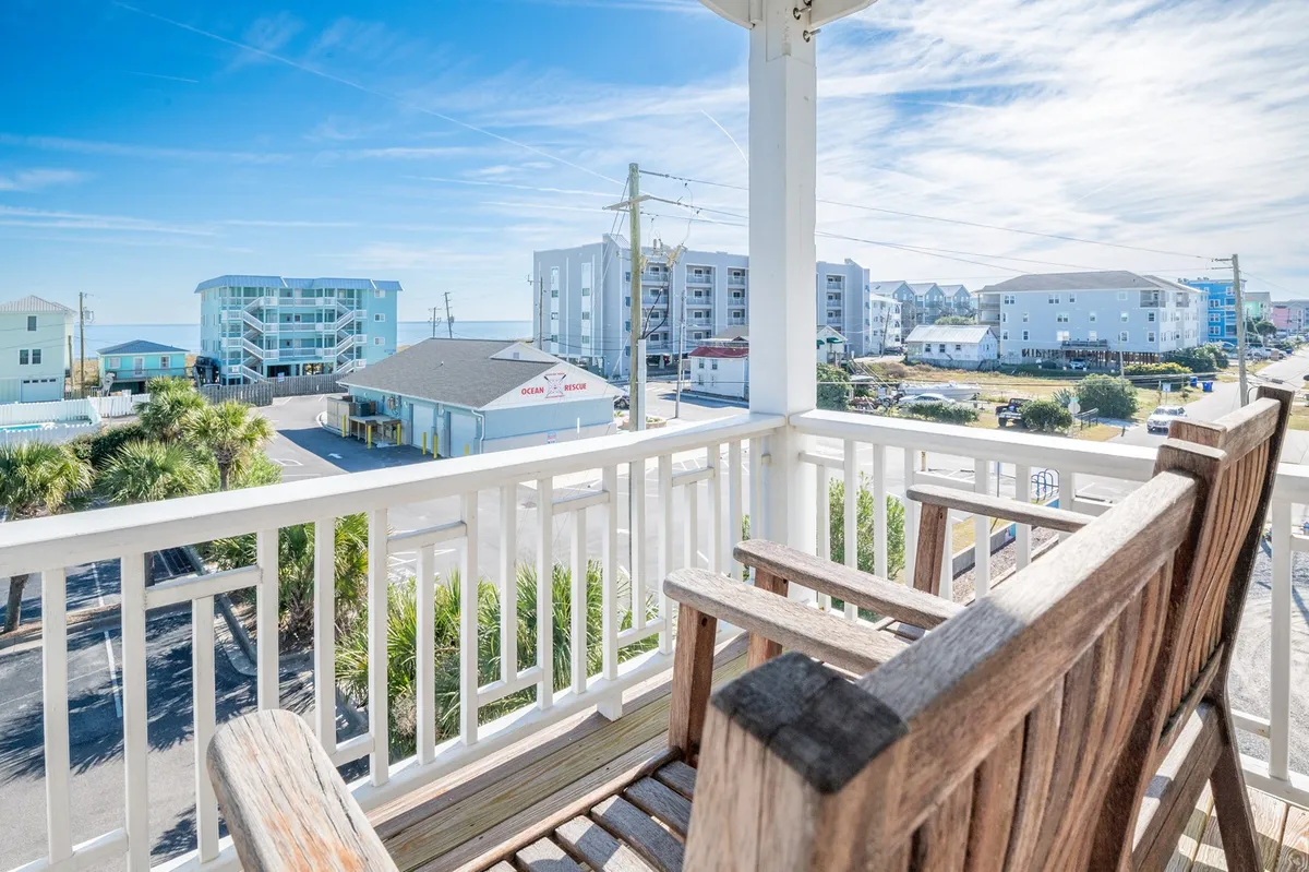 A wooden rocking chair positioned on a porch or balcony with expansive ocean views, palm trees, and surrounding buildings in the distance. The space is ideal for relaxing, reading, or soaking in the coastal breeze, blurring the line between indoor comfort and outdoor beauty.
