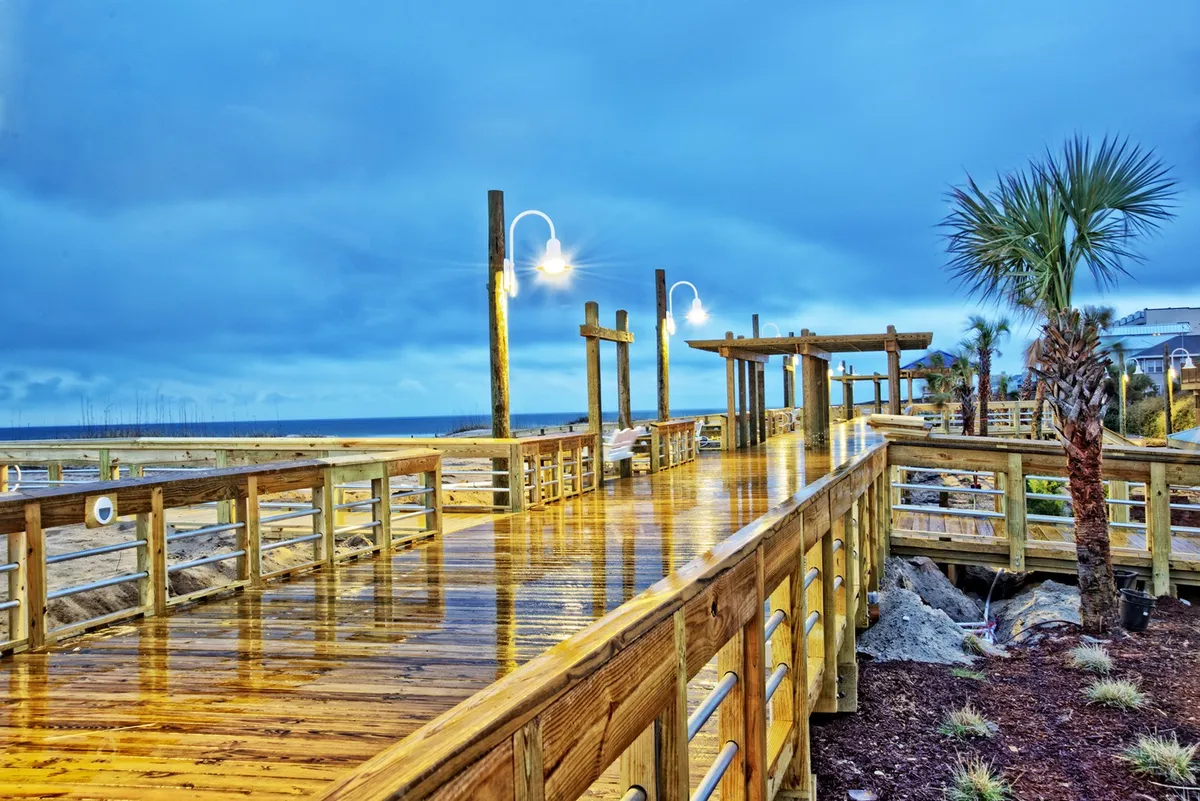 A serene, sunlit wooden boardwalk flanked by palm trees and lampposts, leading visitors toward a beautiful ocean view. The tranquil mood suggests leisurely strolls and the charm of the area’s walkability, with opportunities for both relaxation and exploration.