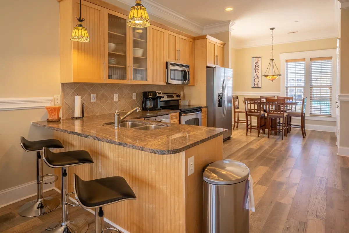 A bright, open kitchen and dining space with a kitchen island, stainless steel appliances, a dining table for six, and beach-themed wall decor. The layout seamlessly connects cooking and dining areas, perfect for family meals and entertaining guests.