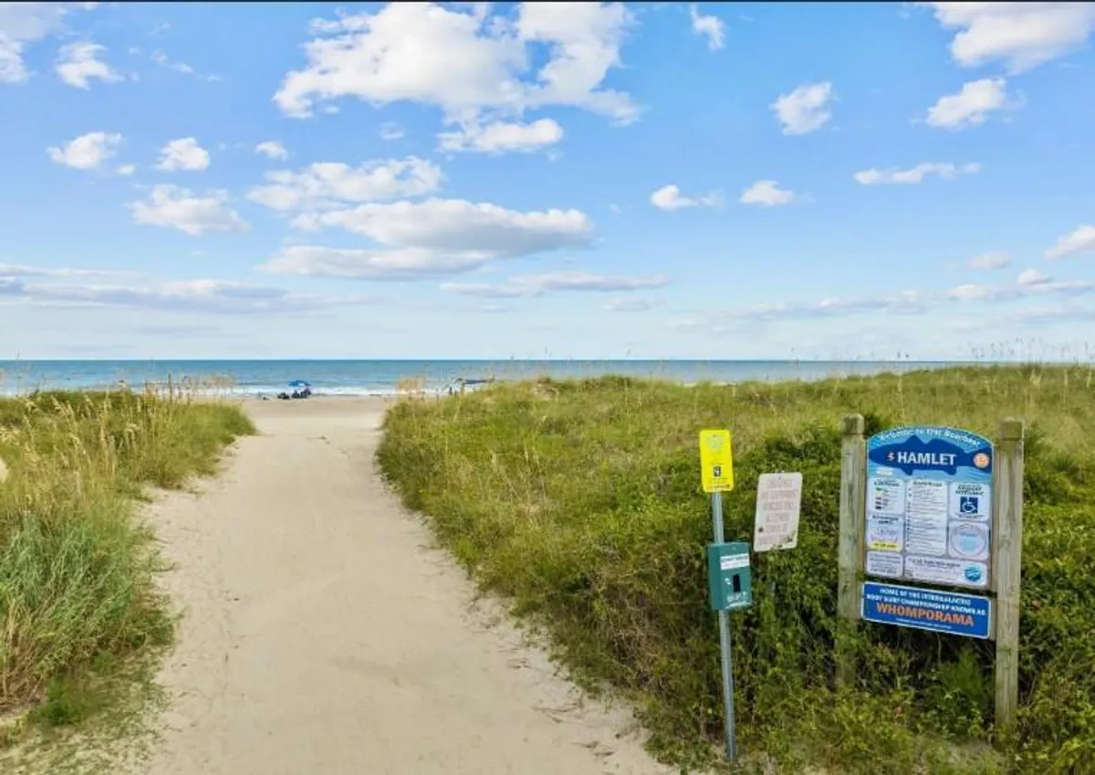 A relaxed beach pathway leads past palm trees and information signposts toward the open ocean, with gentle waves visible in the distance. The tranquil setting hints at easy beach access and leisurely walks.