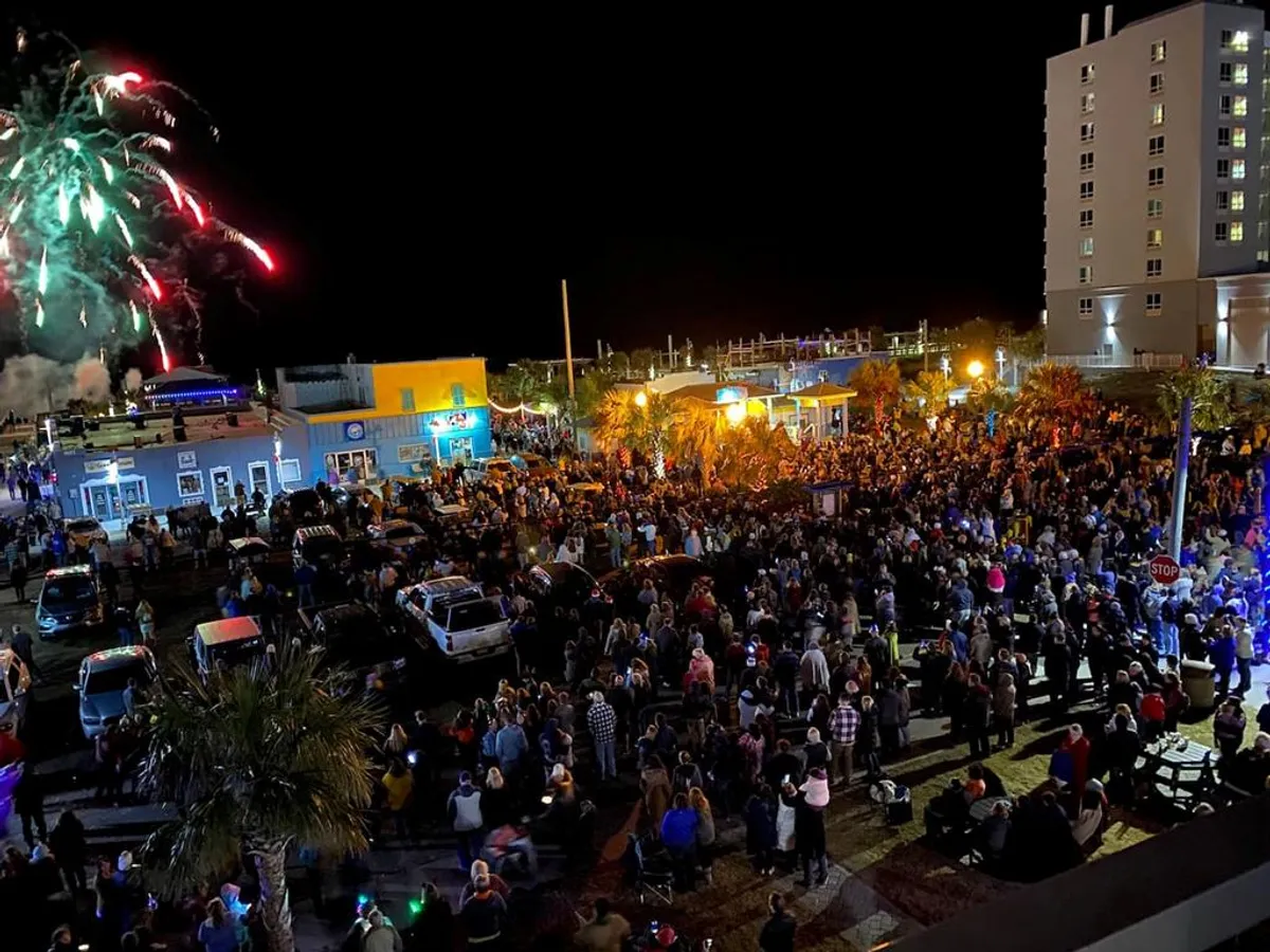 A lively evening scene featuring a crowd gathered to watch fireworks burst over colorful buildings. Palm trees, festive lights, and a joyful crowd capture the celebratory, communal energy of summer fireworks and events on the Boardwalk.