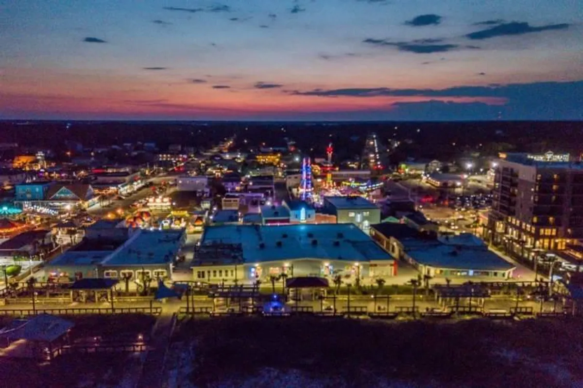 A vibrant nighttime view featuring colorful lights illuminating Carolina Beach's attractions and skyline. The scene includes lively buildings, entertainment venues, and the inviting energy of the boardwalk area, suggesting easy access to exciting local experiences.