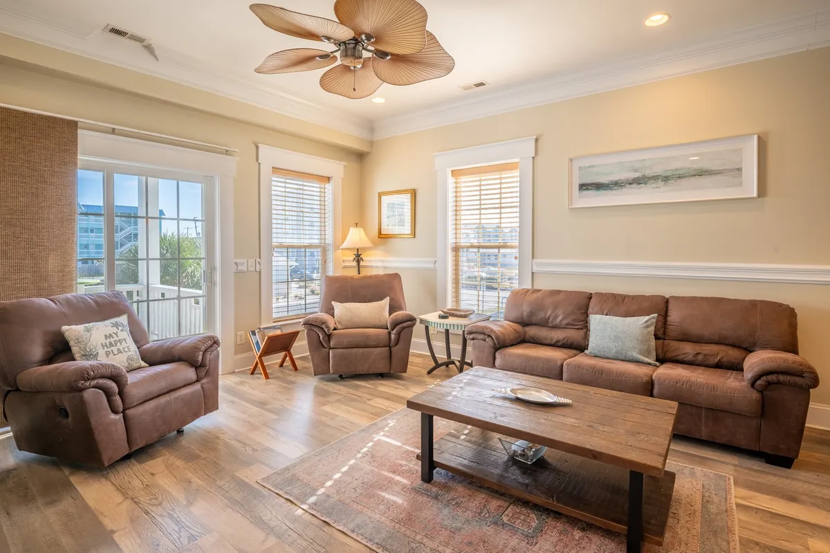A sunlit living room featuring plush leather sofas, a wooden coffee table, a decorative ceiling fan, and artwork on the wall. The arrangement encourages relaxation, socializing, and cozy evenings, with light streaming in to create an inviting atmosphere.
